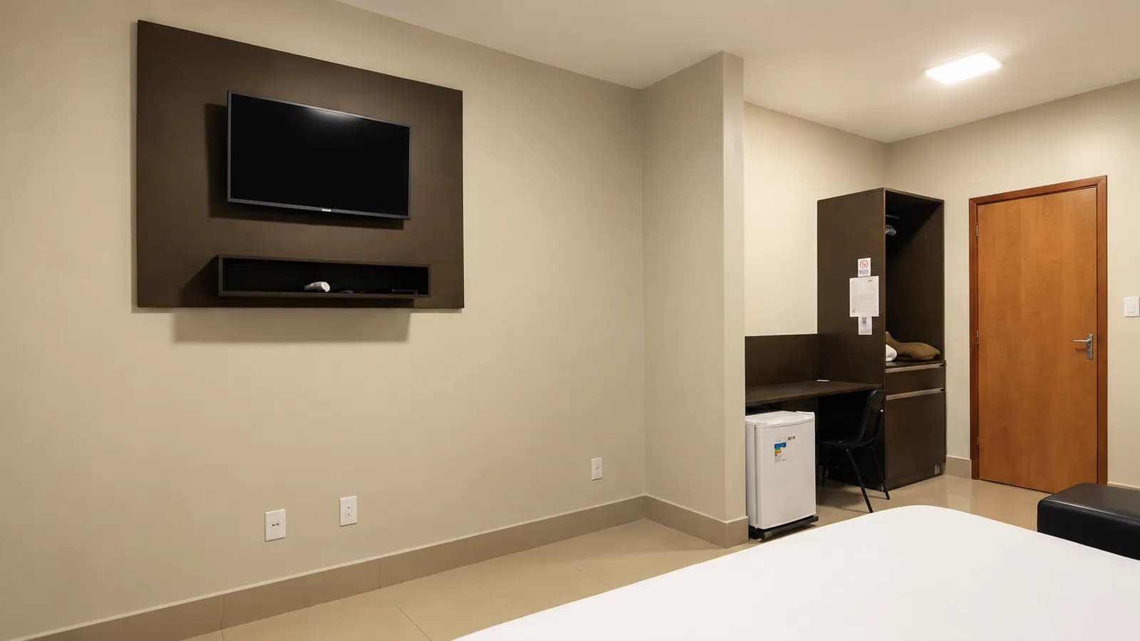 Quarto com televisão na parede, mesa de trabalho, armário e frigobar